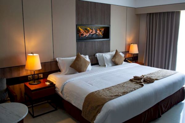 The image depicts a well-lit hotel room with a large bed, two bedside tables with lamps, wall-mounted artwork, and a window with curtains.