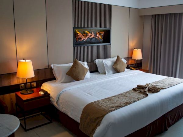 The image depicts a well-lit hotel room with a large bed, two bedside tables with lamps, wall-mounted artwork, and a window with curtains.