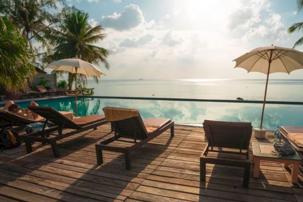 A serene poolside scene with lounge chairs and umbrellas overlooking a calm body of water, under a partly cloudy sky and surrounded by palm trees.
