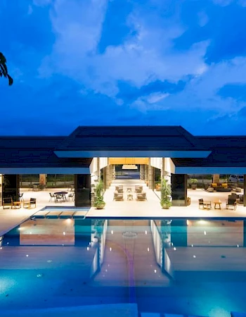 The image shows a luxurious, well-lit modern building with a large swimming pool in the foreground under a blue, twilight sky.