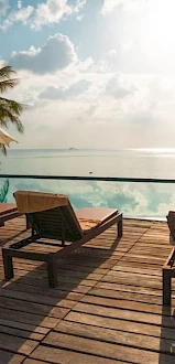 A serene poolside scene with lounge chairs, umbrellas, and palm trees, overlooking a calm ocean under a sunlit sky.