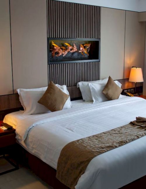 A well-lit hotel room with a large bed, two bedside tables, and lamps. Decor includes a wall-mounted art piece and neatly arranged pillows and linens.