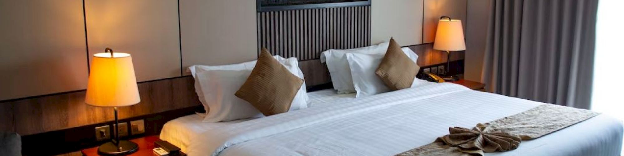 A cozy hotel room with a large bed, lit lamps on both sides, decorative pillows, and a wall-mounted art piece above the headboard.
