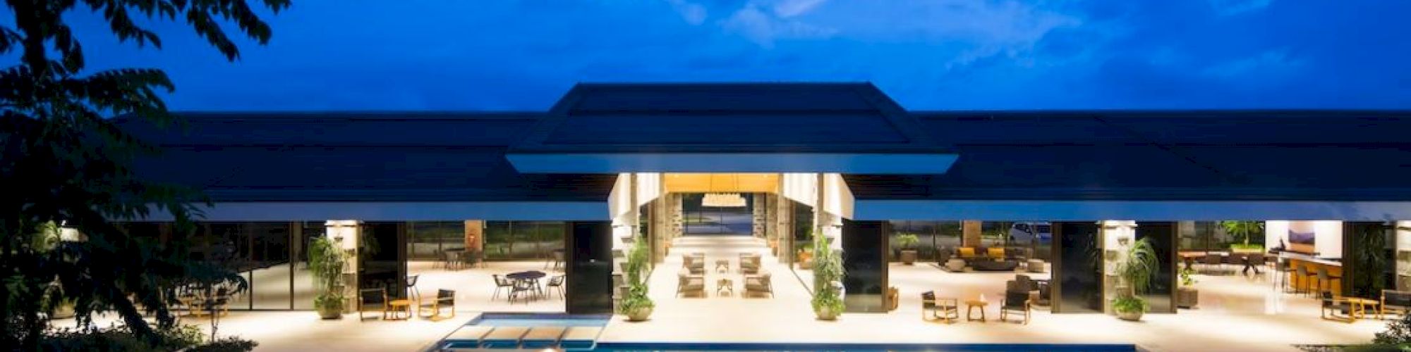 A luxurious open-air building with a large swimming pool in the foreground, modern design, illuminated at dusk under a partly cloudy sky.
