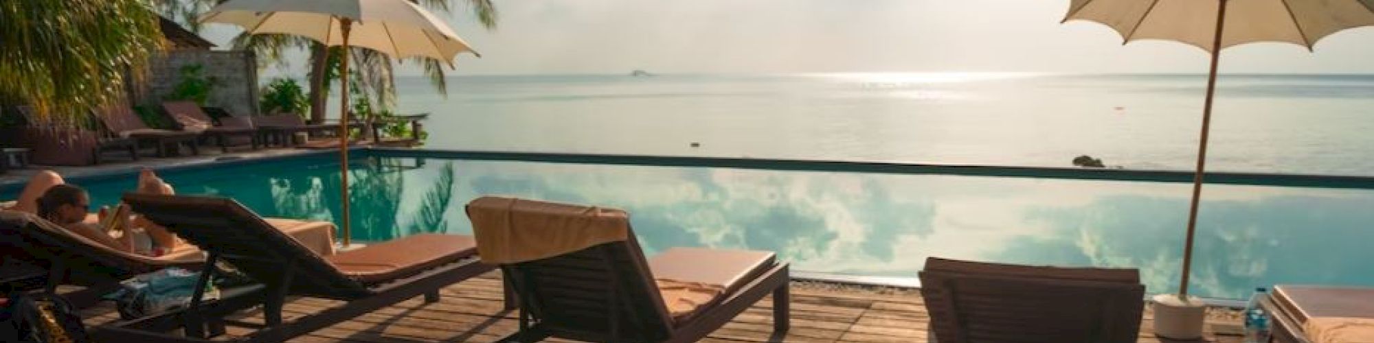 A serene poolside setting with lounge chairs and umbrellas overlooking a calm body of water, surrounded by palm trees and lush greenery.