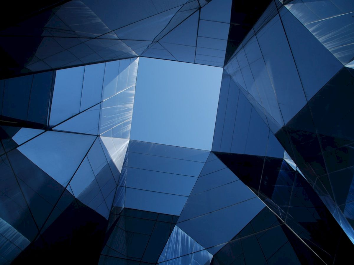 This image depicts a geometric architectural structure made of reflective glass panels, forming an open square space with a view of the sky.