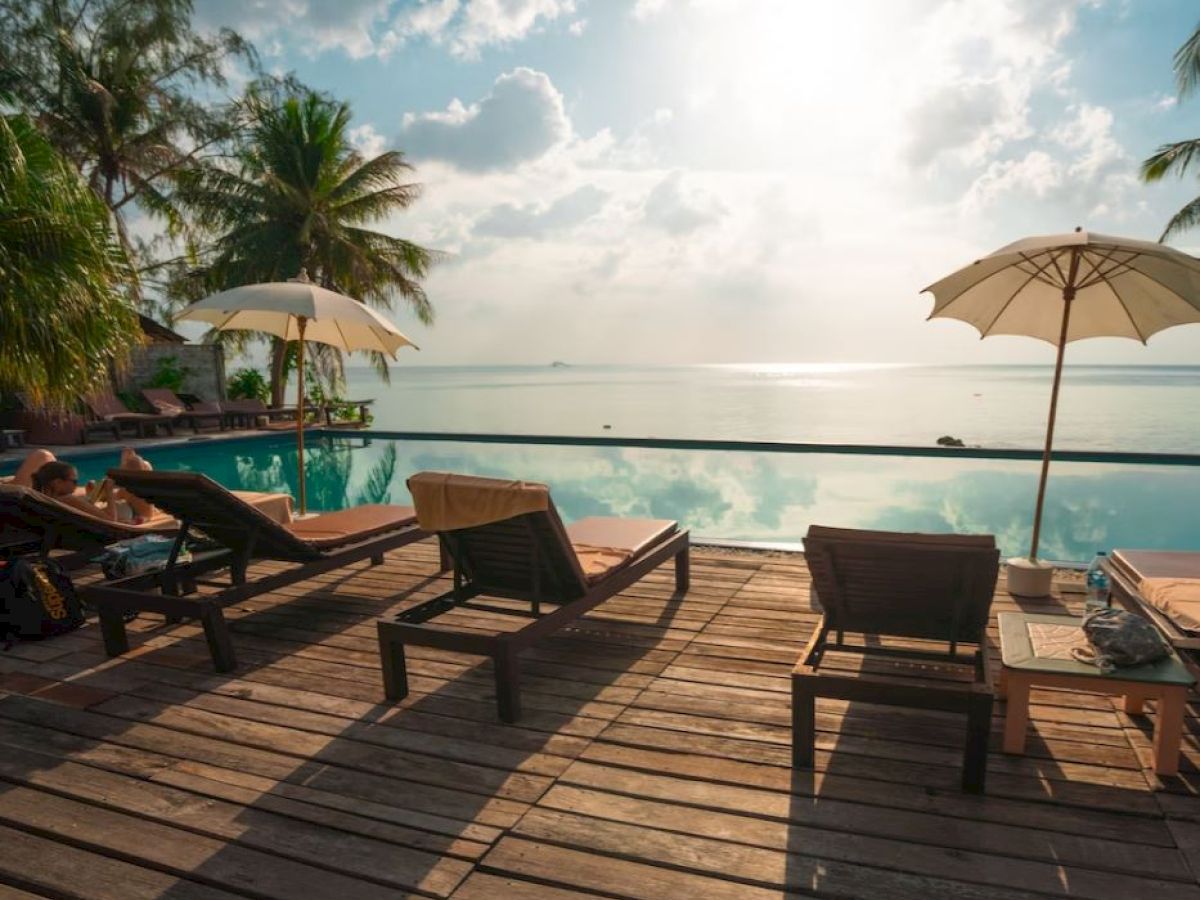 A serene poolside scene with lounge chairs, umbrellas, palm trees, and a view of the ocean under a bright, cloudy sky.