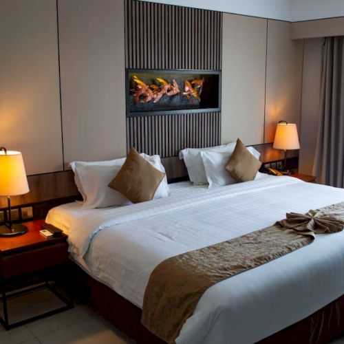 A cozy hotel room with a large bed, pillows, bedside lamps, and a wall-mounted fireplace, adorned with neutral tones and modern décor.