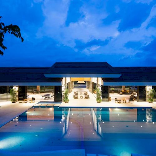 An illuminated luxurious building with a large central swimming pool and surrounding lounge areas at night, with a blue sky backdrop.