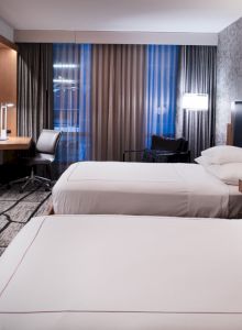 A modern hotel room with two beds, a wall-mounted TV, a work desk, and contemporary furnishings, well-lit with a large window.