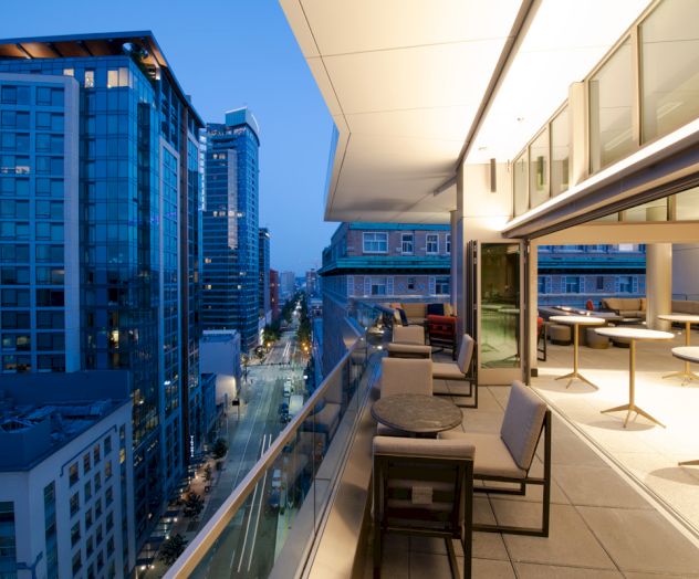 A modern urban balcony at twilight with outdoor seating and tables overlooking a city street filled with tall buildings.