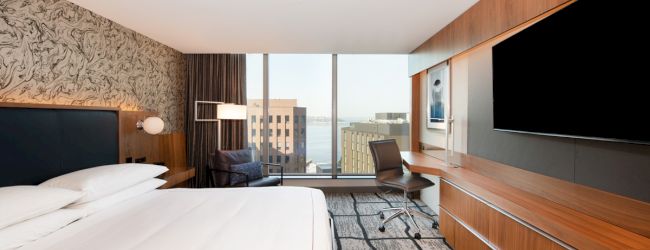 A modern hotel room with a large bed, wall-mounted TV, desk, chair, and window offering a city view.