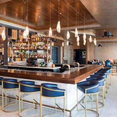 A modern bar with a wooden counter, high chairs, hanging lights, and a fully stocked bar in the background in a sleek interior.