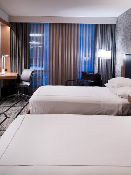The image shows a modern hotel room with two neatly made beds, a large TV, a desk with a chair, and stylish lighting.