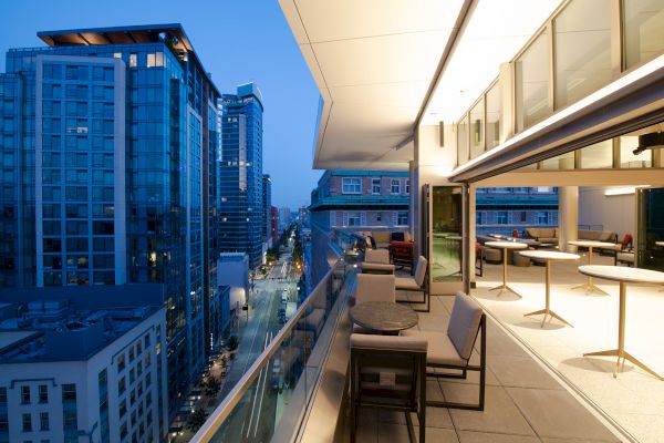 A modern balcony with seating and tables overlooks a cityscape with tall buildings and street lights at dusk, creating a sophisticated ambiance.