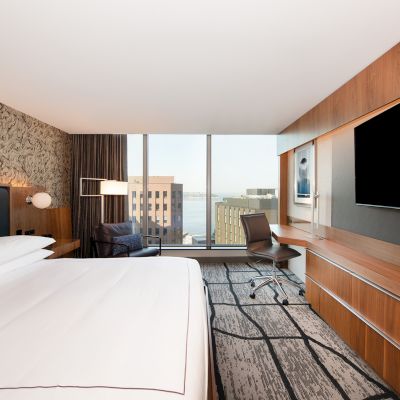 A modern hotel room features a large bed, desk, TV, chair, and a large window with a cityscape view.