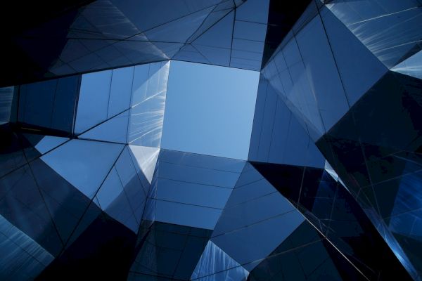 The image shows a modern architectural structure with reflective glass surfaces forming a geometric pattern, revealing a blue sky in the center.