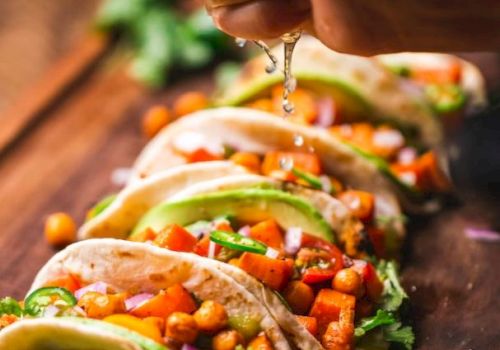 The image shows a person squeezing lime juice over a row of chickpea and vegetable tacos, garnished with cilantro and avocado slices, on a wooden surface.