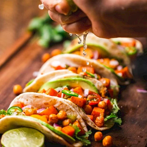 The image shows a person squeezing lime juice over a row of chickpea and vegetable tacos, garnished with cilantro and avocado slices, on a wooden surface.