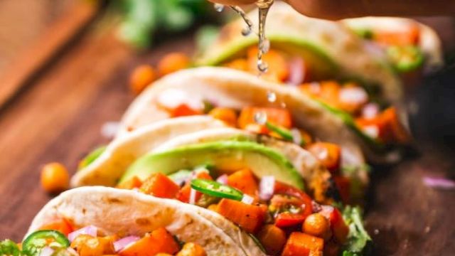 The image shows a person squeezing lime juice over a row of chickpea and vegetable tacos, garnished with cilantro and avocado slices, on a wooden surface.