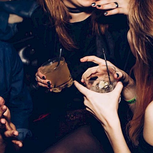People are socializing, laughing, and holding drinks in a dimly lit setting, possibly a bar or a party, with a lively atmosphere.