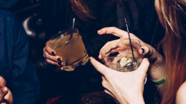 People are socializing, laughing, and holding drinks in a dimly lit setting, possibly a bar or a party, with a lively atmosphere.