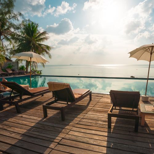 A serene beachfront scene featuring lounge chairs, umbrellas, a wooden deck, and an infinity pool overlooking a calm sea under a sunny sky.