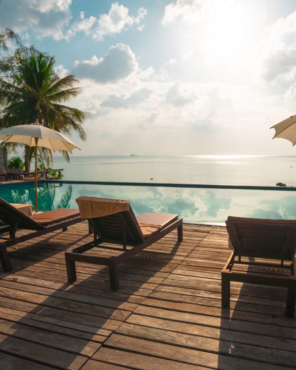 A serene beachfront scene featuring lounge chairs, umbrellas, a wooden deck, and an infinity pool overlooking a calm sea under a sunny sky.