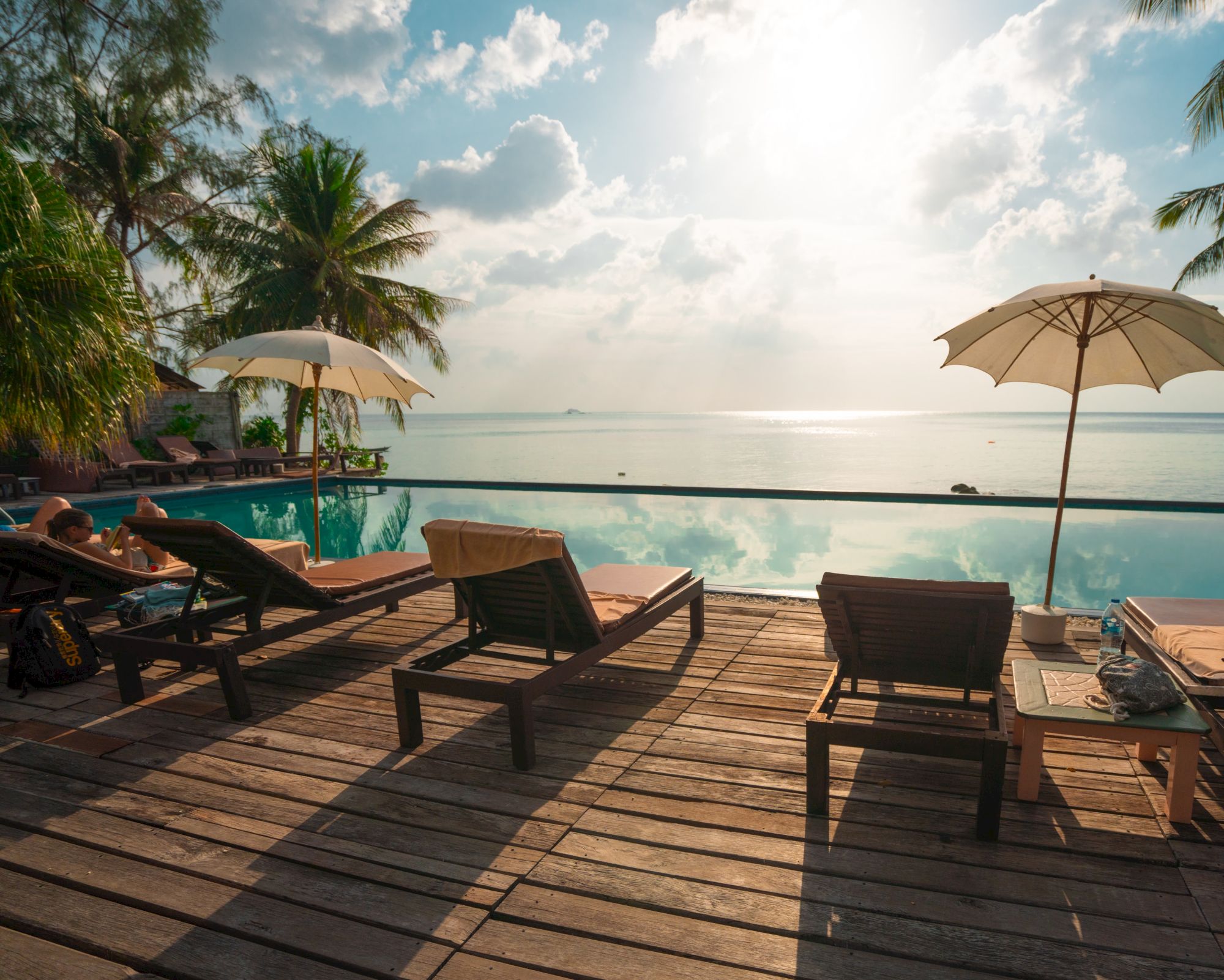 Sun loungers and umbrellas beside a pool with palm trees and an ocean view, under a partly cloudy sky with the sun shining down.