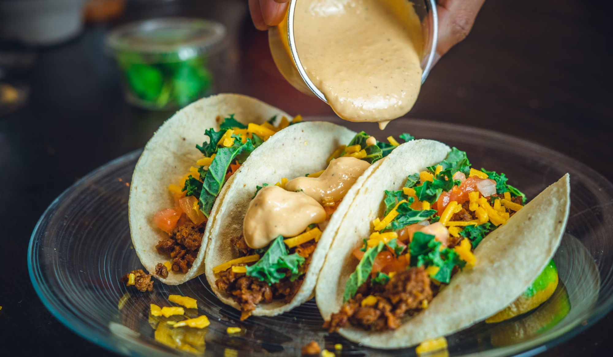 Three tacos on a plate, topped with lettuce, tomatoes, cheese, and sauce being poured over them from a small container.