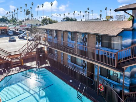 The image shows a motel with a central pool, a two-tiered building, and nearby parked cars. In the distance, there's a row of palm trees and shops.