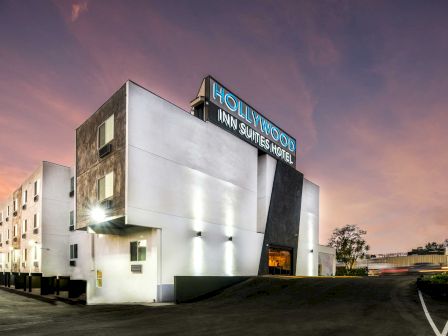 This image shows the exterior of a Hollywood Inn & Suites hotel building during sunset or dusk. The sky is colorful with a blend of pink and purple hues.