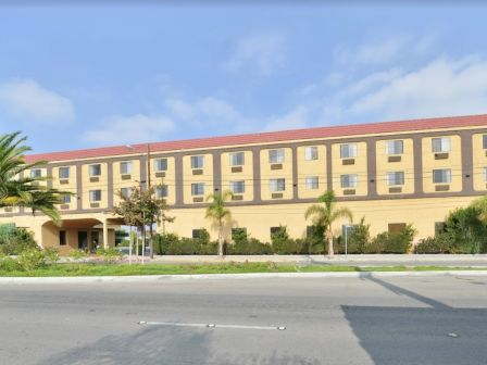 A beige, four-story building with a red roof, palm trees, and shrubs in front, situated beside a road under a clear sky.