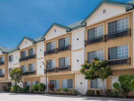 The image shows the exterior of a three-story building with multiple windows and balconies, surrounded by greenery and shrubs.