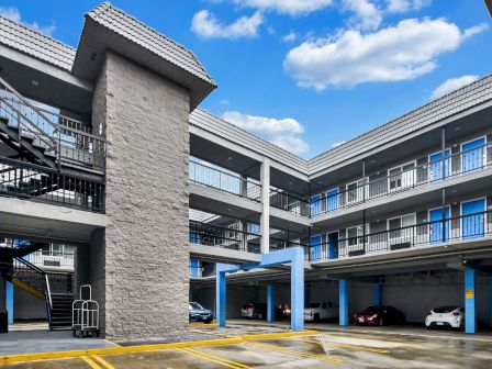 This image shows an exterior view of a multi-story building with a parking area, stairs, and cars parked below. The sky is partly cloudy.