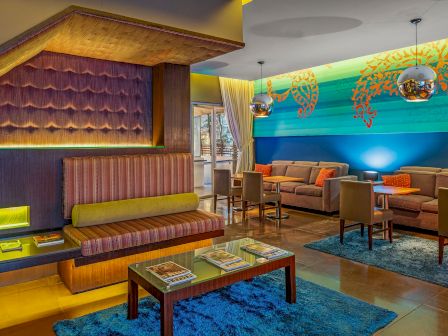 A modern lounge with colorful decor, featuring couches, tables, pendant lights, and a blue rug, creating a vibrant, inviting atmosphere.