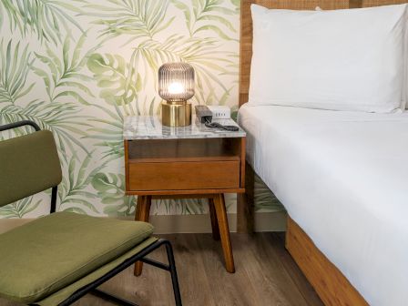 A stylish bedroom with a leafy wallpaper, a wooden bedside table with a lamp, and a green chair on wooden flooring next to a bed.