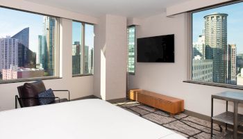The image depicts a modern bedroom with large windows offering a cityscape view, a wall-mounted TV, a chair, and minimalistic decor.