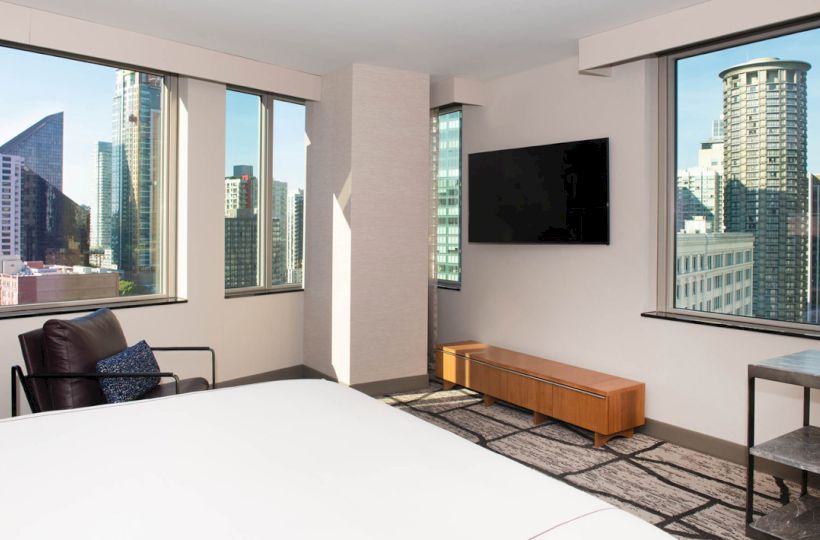 The image depicts a modern bedroom with large windows offering a cityscape view, a wall-mounted TV, a chair, and minimalistic decor.