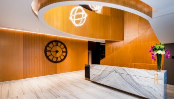 The image shows a modern lobby with a marble reception desk, a large wall clock, wooden interior, stylish lighting fixtures, and a vase of flowers.