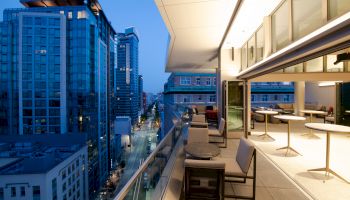 A modern rooftop terrace at dusk with outdoor seating overlooks tall city buildings and a busy street below, illuminated by streetlights.