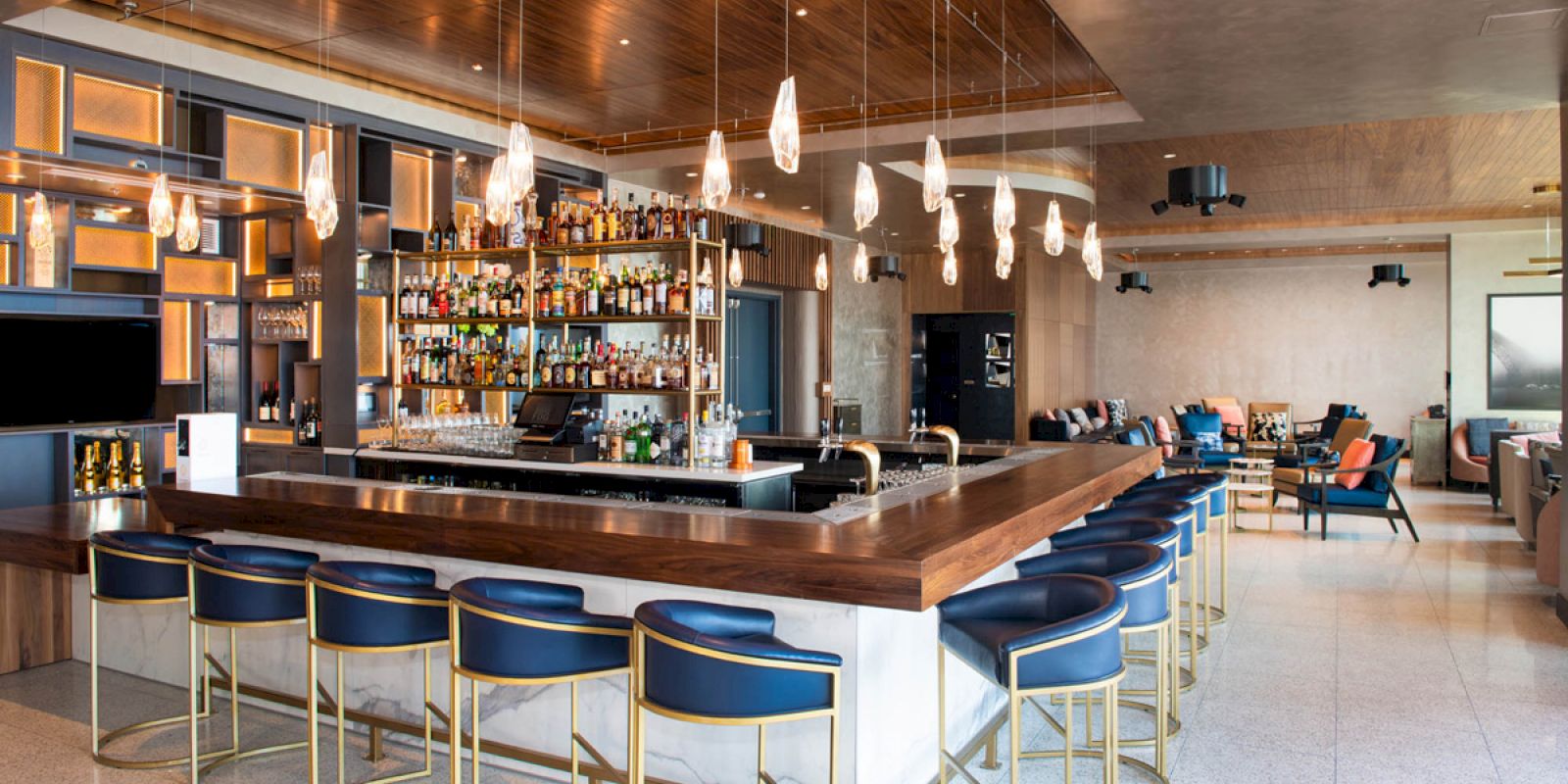 A modern bar with blue high-back chairs, a well-stocked liquor display, pendant lighting, and seating areas in the background.