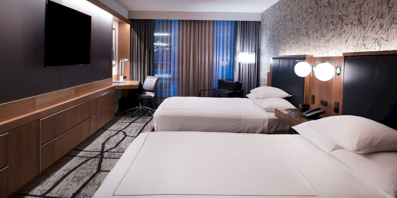 The image shows a modern hotel room with two double beds, a wall-mounted TV, a desk, and large windows with curtains.