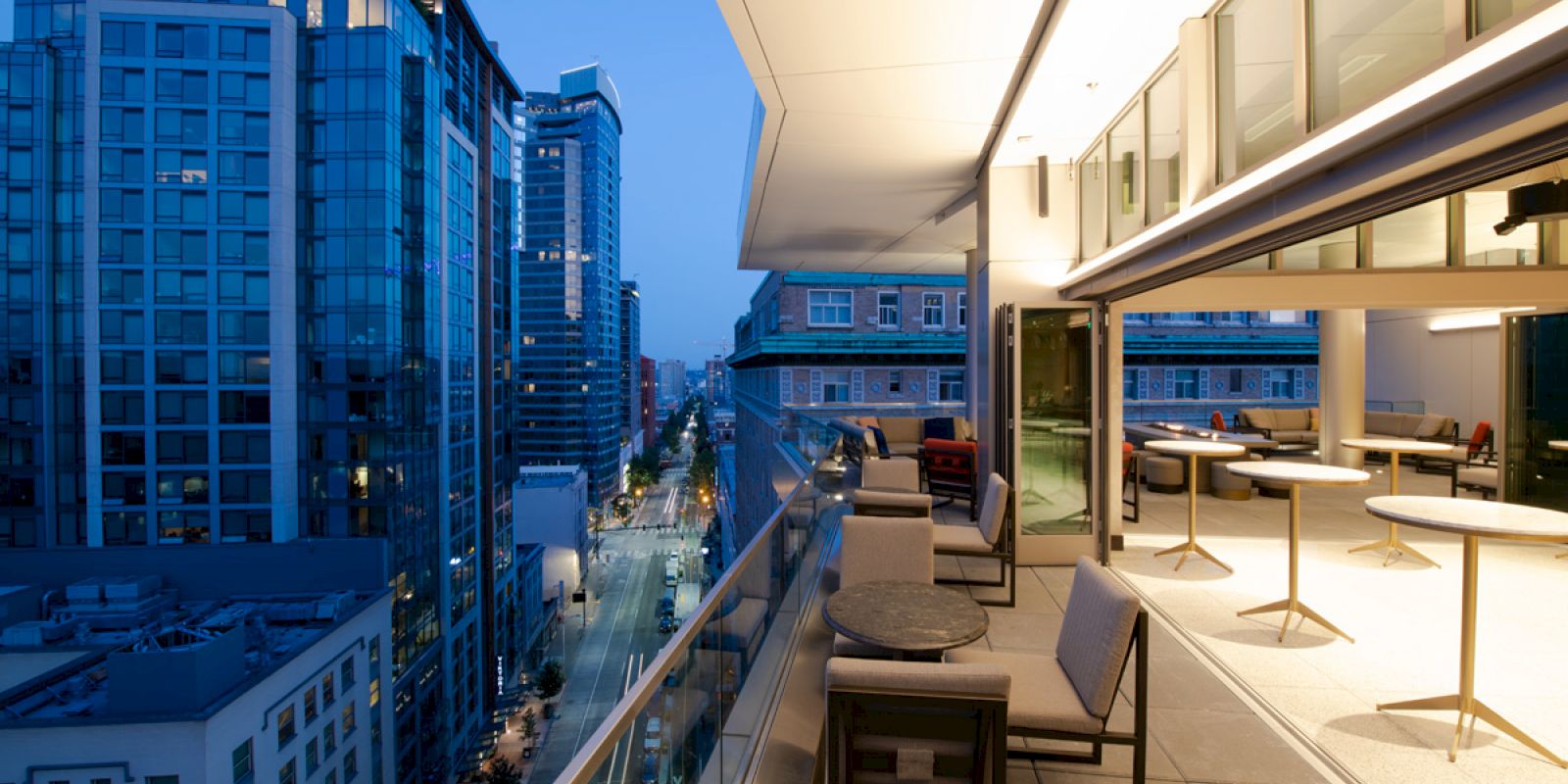 A modern balcony overlooks a cityscape with tall buildings at dusk. The balcony has tables and chairs, and the inside area is visible through glass walls.