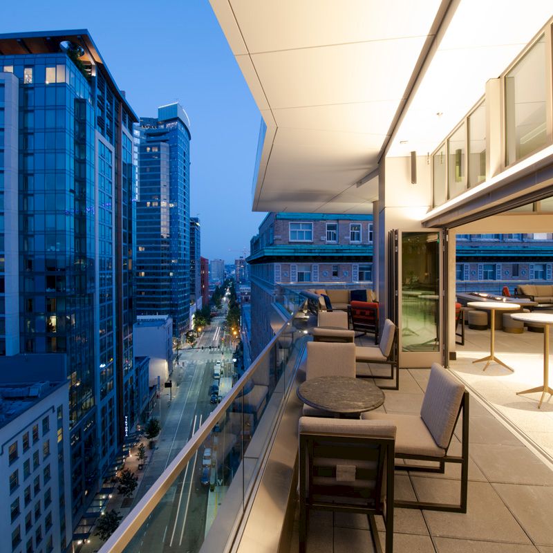 A modern rooftop terrace overlooking a cityscape at dusk, with outdoor seating, tables, and indoor spaces visible through large windows.