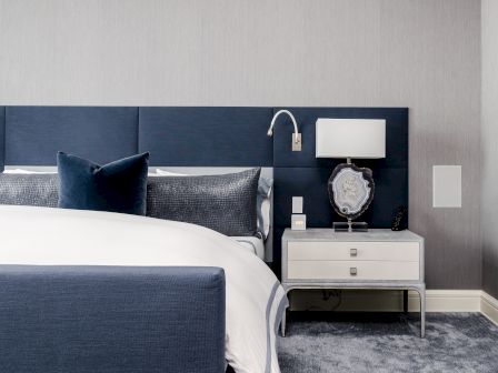 A modern bedroom features a blue headboard, white bed linens, navy pillows, a bedside table with a lamp, and an artistic décor piece.