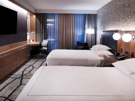 A modern hotel room features two neatly made beds, a wall-mounted TV, a work desk with a chair, and stylish lighting and decor.