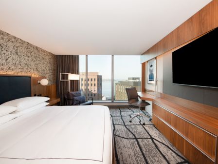A modern hotel room featuring a large bed, desk, chair, TV, and a view of the city through a large window, with stylish decor.