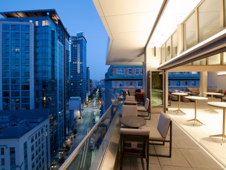 The image shows a modern balcony with seating, overlooking a cityscape with tall buildings and a street at dusk.