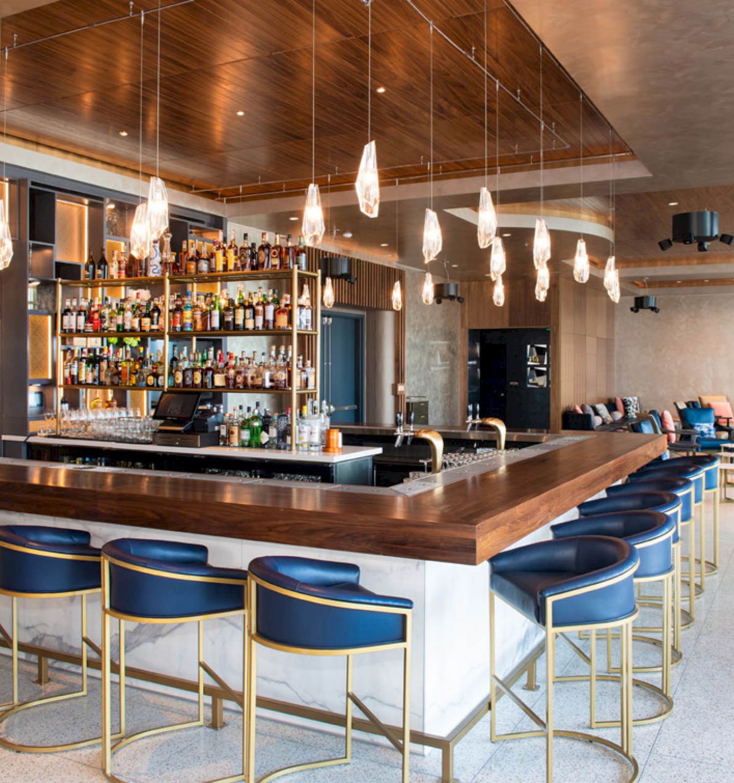 A modern bar with blue stools, a well-stocked counter, hanging lights, and a cozy seating area in the background, blending style and comfort.